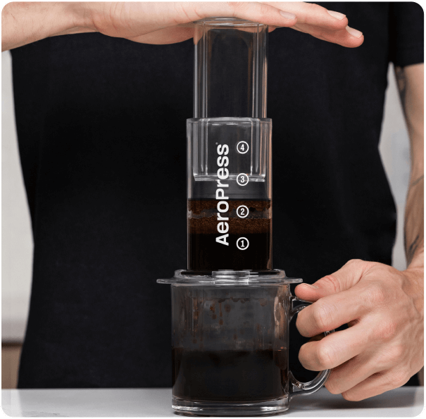 Transparent AeroPress coffee brewer in use, hands pressing the plunger to brew coffee
