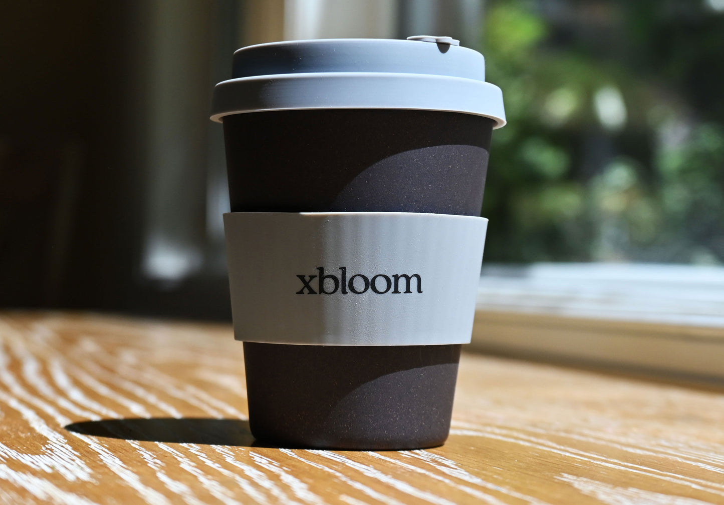 Reusable xBloom coffee cup with a textured dark body and light gray lid, lit by natural sunlight on a wooden surface