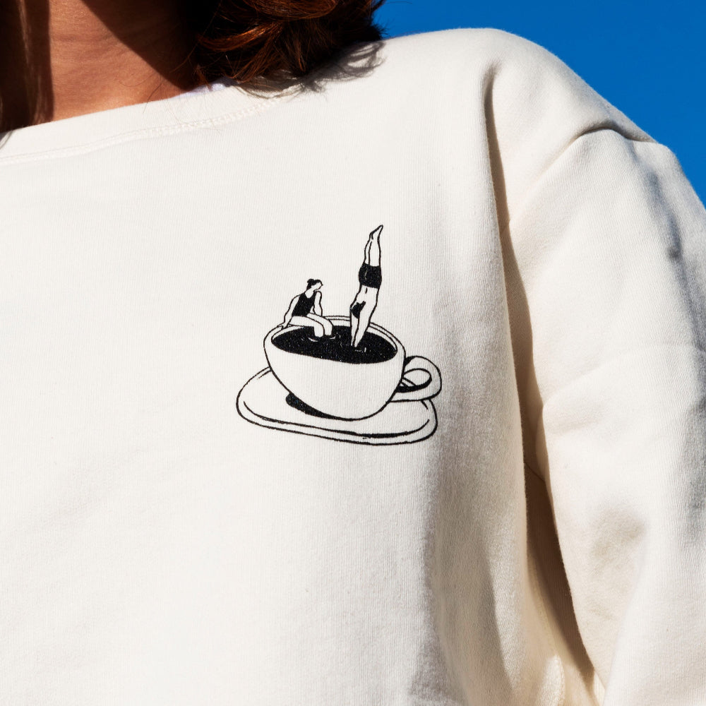 Person wearing a white sweatshirt with a black coffee cup design on the back against a blue sky.