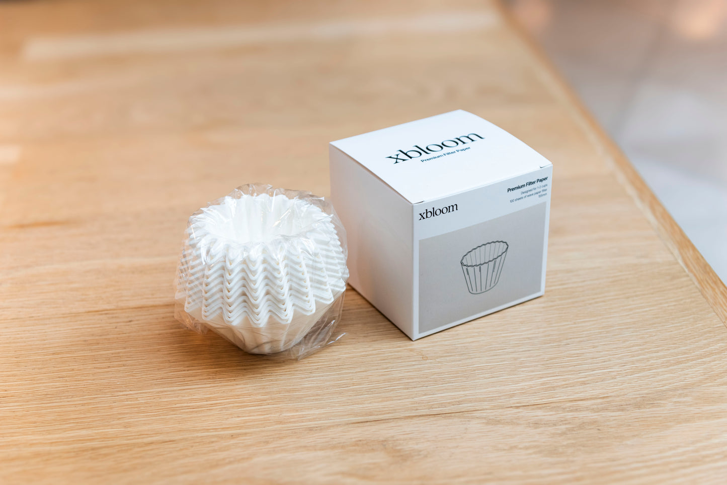 Stack of xBloom premium pleated coffee filters in clear wrap, shown next to the branded white product box on a light wood surface.