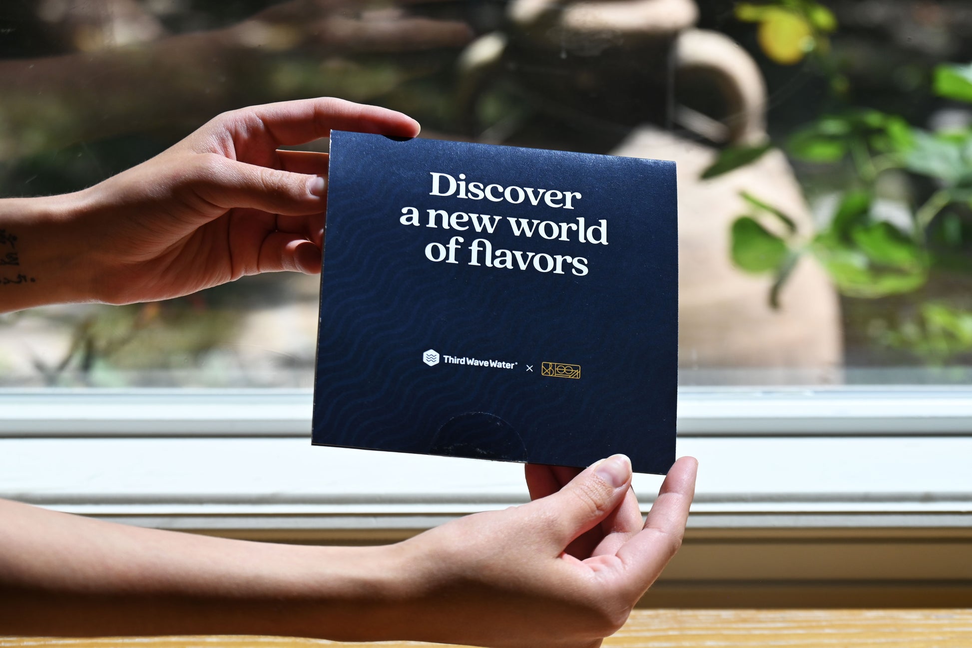 Hands holding a dark blue Third Wave Water x xBloom co-branded card that reads 'Discover a new world of flavors', in front of a bright window