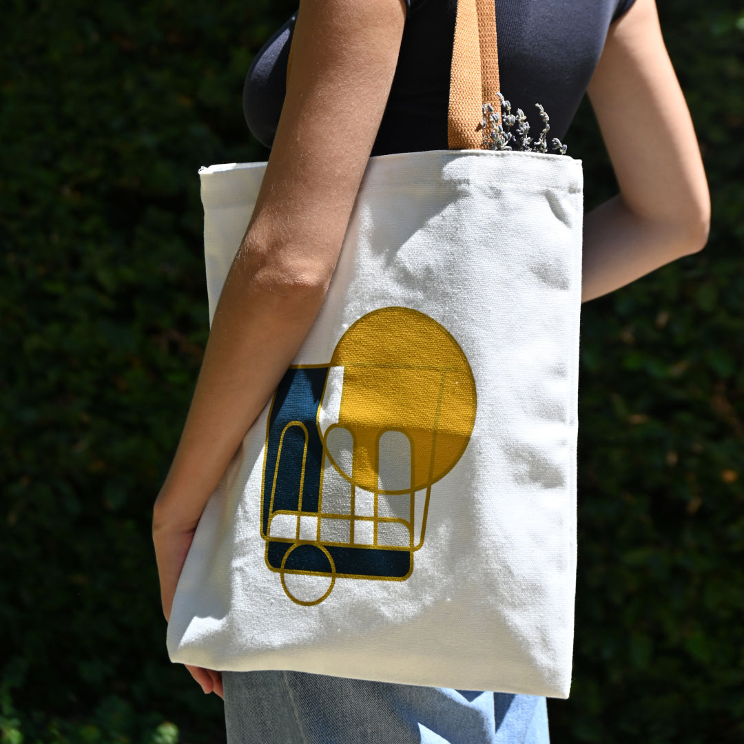 Person carrying a white canvas tote bag with tan handles, featuring a modern geometric coffee cup print in navy and gold, outdoors against green foliage