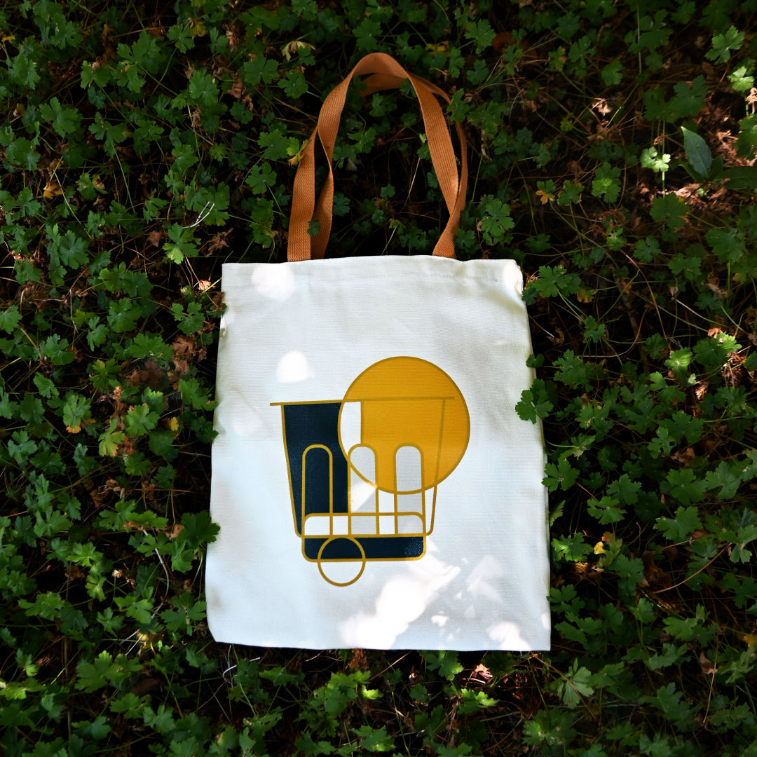 White canvas tote bag with tan handles, featuring a geometric coffee cup graphic in navy and gold, laid flat on green foliage
