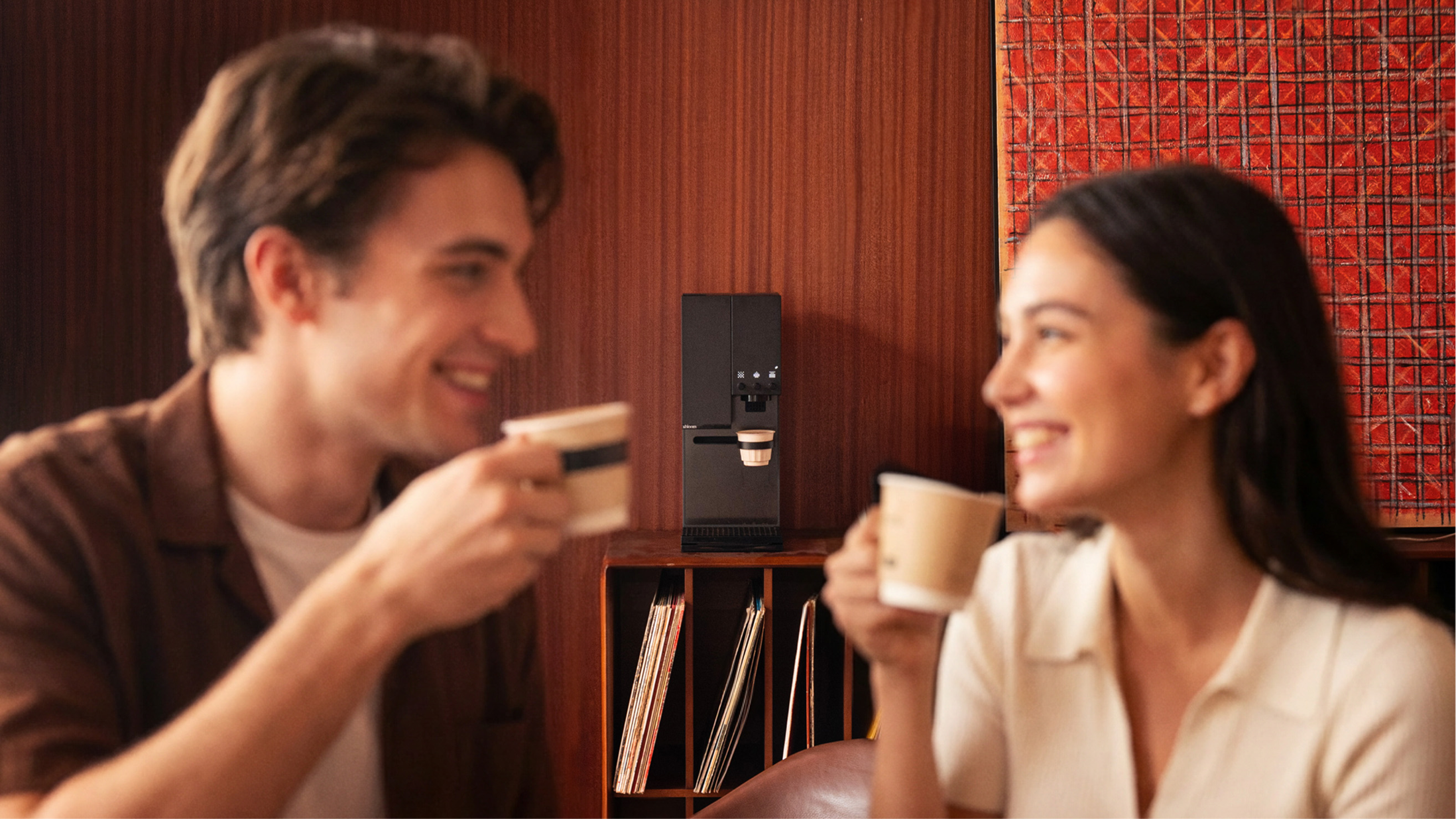 Man and woman sitting together, each holding a coffee cup, with xBloom Studio in the background.